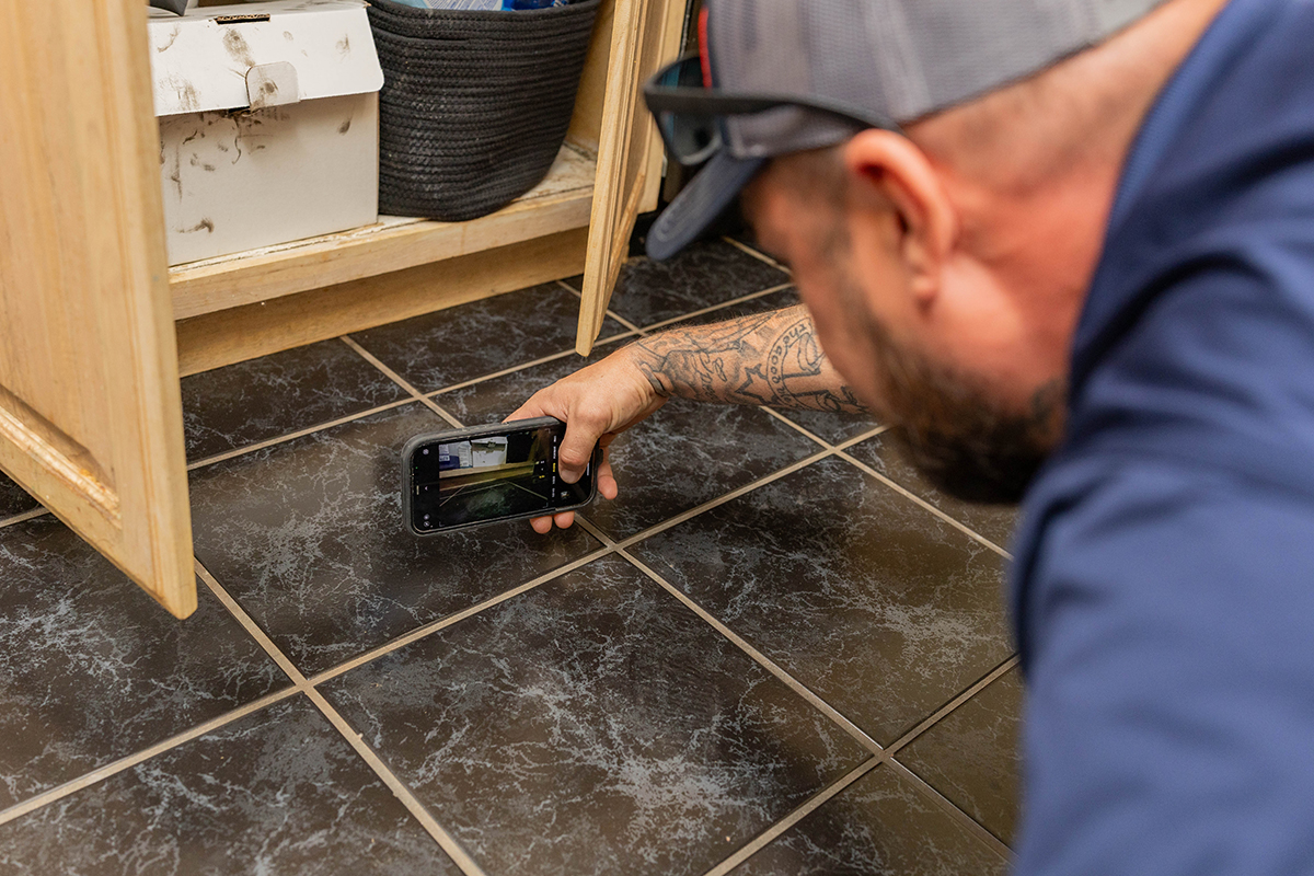 Technician documenting hidden damage during mold removal by inspecting beneath cabinetry in a residential home